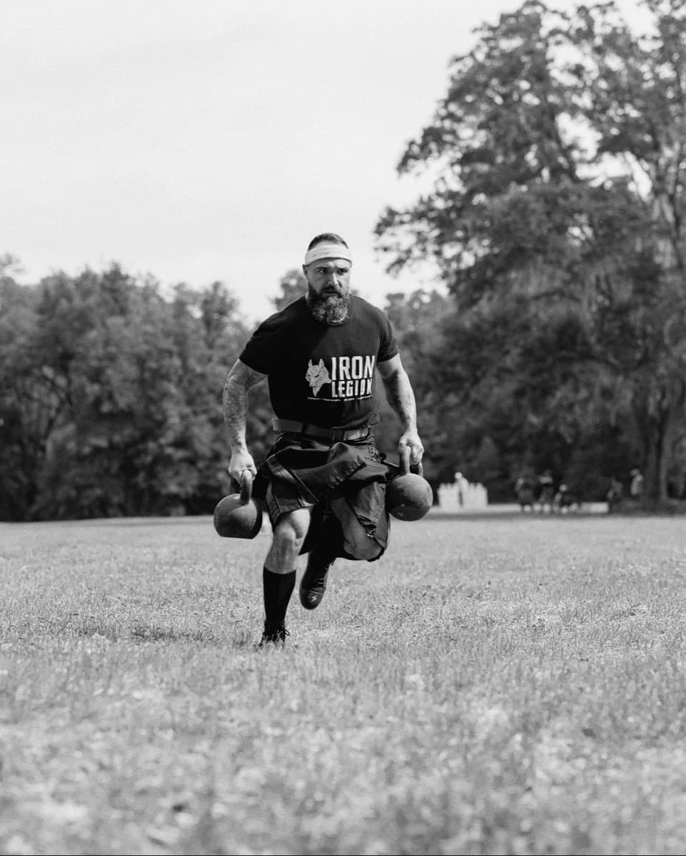 Sean running a loaded carry across a field in an Iron Legion shirt.