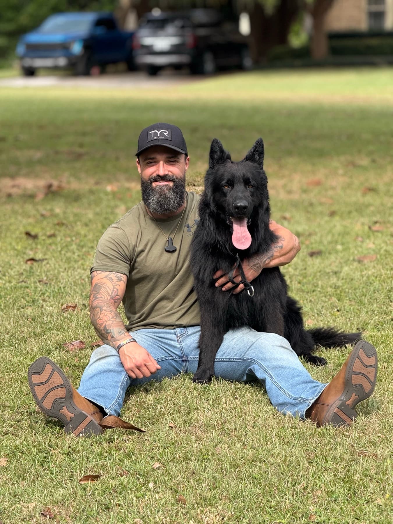 Sean Riley sitting on the grass with his black German Shepherd.
